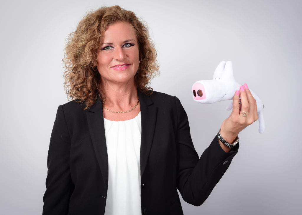 Lächelnde Frau im Anzug hält ein Plüsch-Schwein. Fröhliche, positive Ausstrahlung.